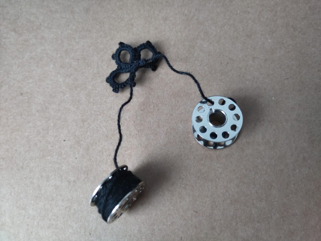 Eine angefangene Occispitze, bei der das Garn auf Nähmaschinenspulen statt Occhischiffchen gewickelt ist.