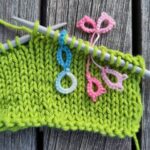 Die Maschenmarkierer im Einsatz. Ein hellblauer Maschenmarkierer steckt auf einer Nadel mit einem angefangenem Strickstück. Ein rosa, schliefenförmiger Maschenmarkierer steckt zwischen zwei Maschen.