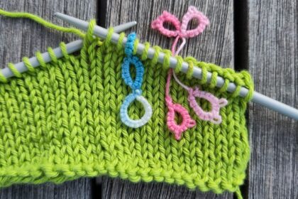 Die Maschenmarkierer im Einsatz. Ein hellblauer Maschenmarkierer steckt auf einer Nadel mit einem angefangenem Strickstück. Ein rosa, schliefenförmiger Maschenmarkierer steckt zwischen zwei Maschen.