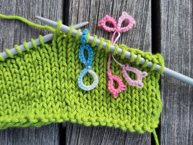 Die Maschenmarkierer im Einsatz. Ein hellblauer Maschenmarkierer steckt auf einer Nadel mit einem angefangenem Strickstück. Ein rosa, schliefenförmiger Maschenmarkierer steckt zwischen zwei Maschen.