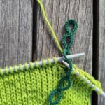 Ein grüner Reihenzähler im Einsatz auf den Stricknadeln. Der Zähler sitzt mit dem dritten Ring auf der linken Stricknadel. Die linke Nadel sticht gerade in den vierten Ring ein.