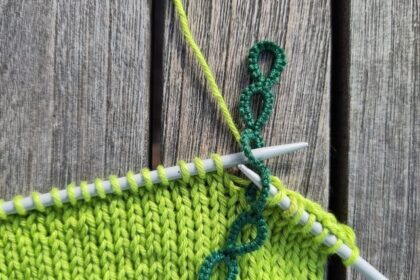 Ein grüner Reihenzähler im Einsatz auf den Stricknadeln. Der Zähler sitzt mit dem dritten Ring auf der linken Stricknadel. Die linke Nadel sticht gerade in den vierten Ring ein.