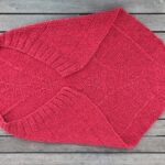 Eine rote Babydecke mit einer fast dreieckigen Form. An der langen Kante ist ein Rippenbund. An dieser Kante wölbt sich die Decke, so dass sie sich besonders gut um ein Baby wickeln lässt.