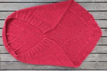 Eine rote Babydecke mit einer fast dreieckigen Form. An der langen Kante ist ein Rippenbund. An dieser Kante wölbt sich die Decke, so dass sie sich besonders gut um ein Baby wickeln lässt.