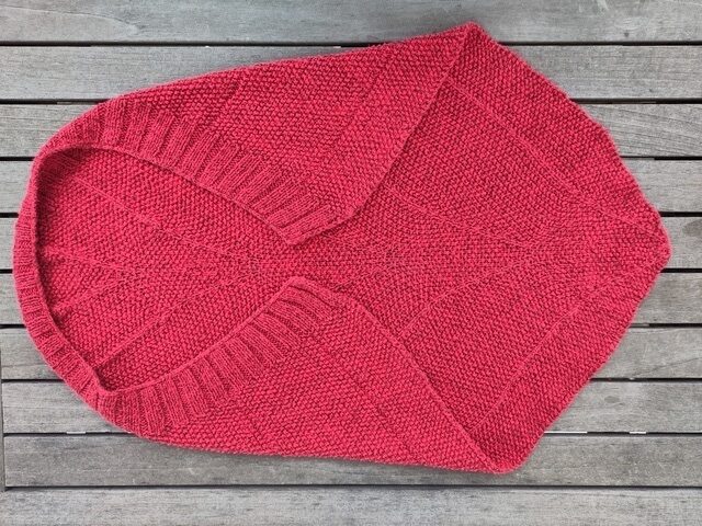 Eine rote Babydecke mit einer fast dreieckigen Form. An der langen Kante ist ein Rippenbund. An dieser Kante wölbt sich die Decke, so dass sie sich besonders gut um ein Baby wickeln lässt.