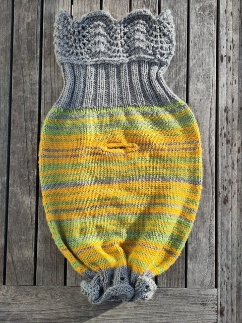 Ein grau, gelb und grün gestreifter Babystrampelsack mit einem Rippenbund und einem zusätzlichen Wellenbund. Der Saum ist zusammen gekräuselt. In der Mitte des Sacks befindet sich eine Öffnung für den Anschnallgurt der Babyschale. Die Form erinnert an einen Fisch.