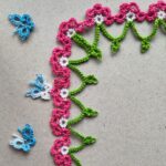 Eine Blumenborte in Occhitechnik mit grünen Bögen und rosa-weißen Blütenelementen. Die Borte hat ein Eckelement. Links im Bild befinden sich drei blaue SChmetterlinge in Occhitechnik.