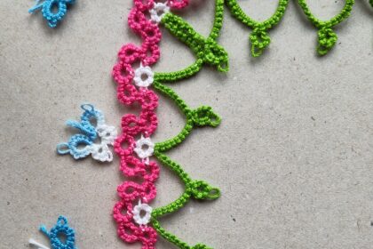 Eine Blumenborte in Occhitechnik mit grünen Bögen und rosa-weißen Blütenelementen. Die Borte hat ein Eckelement. Links im Bild befinden sich drei blaue SChmetterlinge in Occhitechnik.