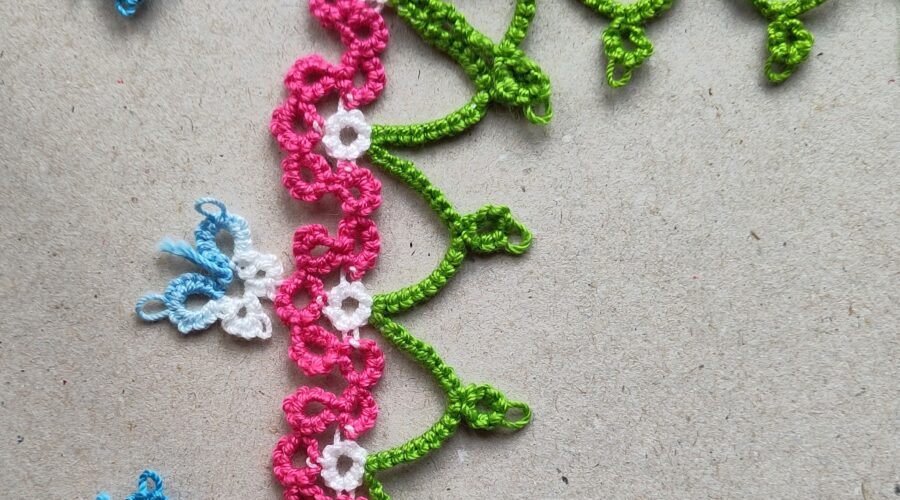 Eine Blumenborte in Occhitechnik mit grünen Bögen und rosa-weißen Blütenelementen. Die Borte hat ein Eckelement. Links im Bild befinden sich drei blaue SChmetterlinge in Occhitechnik.