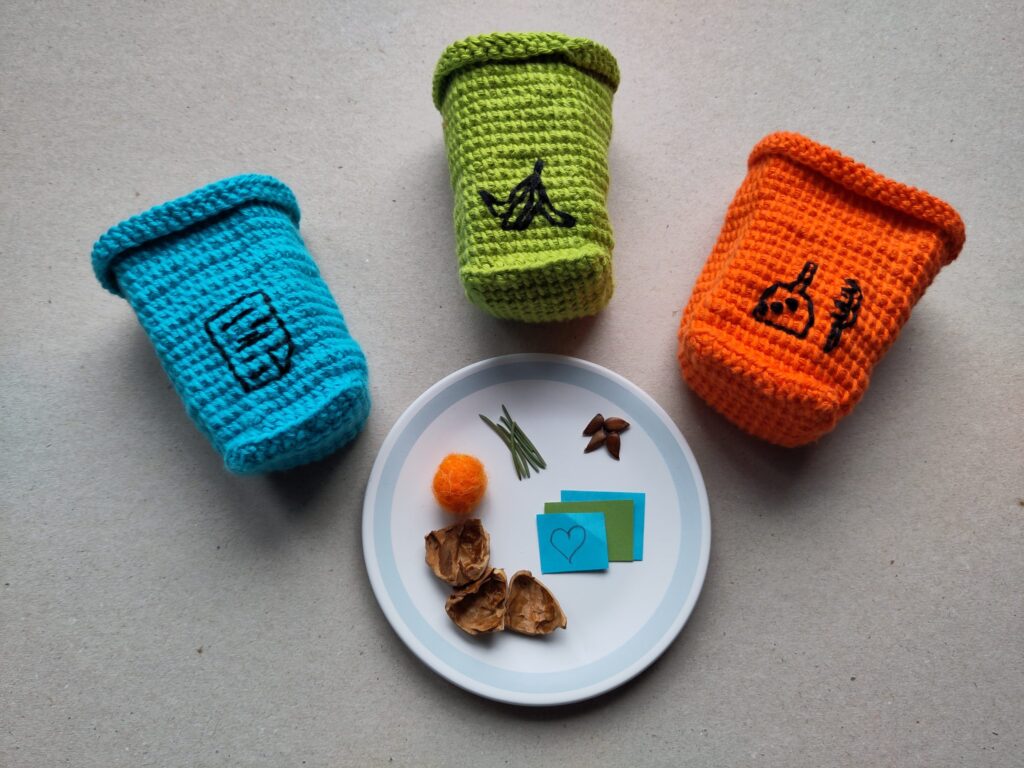 drei Gehäkelte Mülltonnen in blau, grün und orange sind um einen Teller mit Müllteilchen arrangiert.