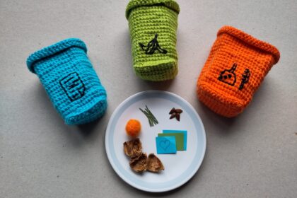 Drei Gehäkelte Mülltonnen in blau, grün und orange sind um einen Teller mit Müllteilchen arrangiert.