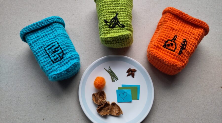 drei Gehäkelte Mülltonnen in blau, grün und orange sind um einen Teller mit Müllteilchen arrangiert.
