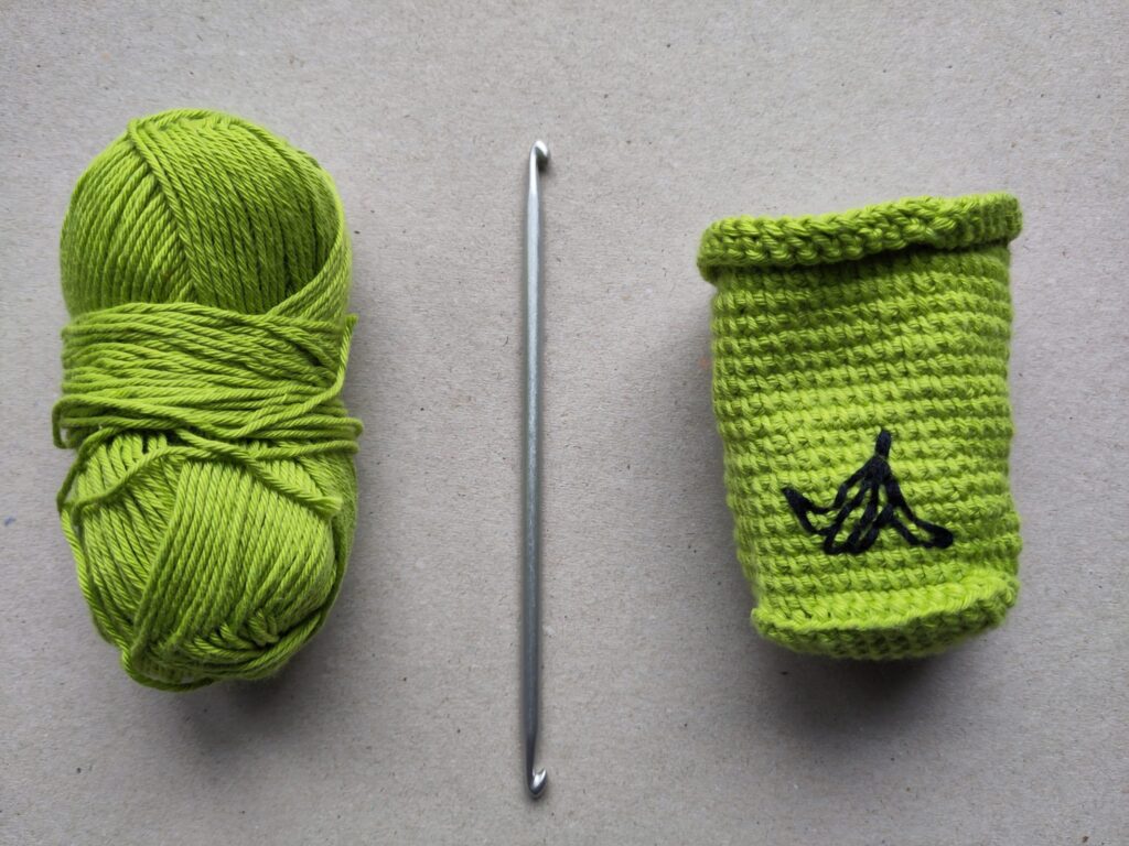 VOn links nach rechts ein günes Wollknäul, eine Tunesische Häkelnadel mit einem Haken an jedem Ende und eine grüne, gehäkelte Mülltonne mit einer aufgestickten Bananenschale.