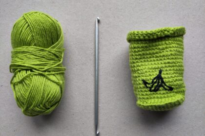 Von links nach rechts ein günes Wollknäul, eine Tunesische Häkelnadel mit einem Haken an jedem Ende und eine grüne, gehäkelte Mülltonne mit einer aufgestickten Bananenschale.