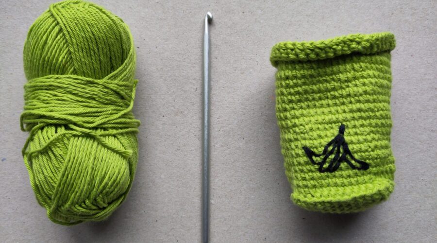 Von links nach rechts ein günes Wollknäul, eine Tunesische Häkelnadel mit einem Haken an jedem Ende und eine grüne, gehäkelte Mülltonne mit einer aufgestickten Bananenschale.