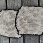 Zwei tunesisch gehäkelte, beigefarbene Glasuntersetzer mit schwarzem Rand auf einem Holztisch