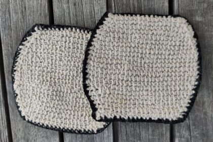 Zwei tunesisch gehäkelte, beigefarbene Glasuntersetzer mit schwarzem Rand auf einem Holztisch