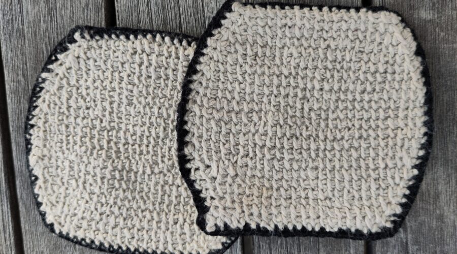 Zwei tunesisch gehäkelte, beigefarbene Glasuntersetzer mit schwarzem Rand auf einem Holztisch