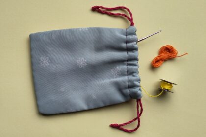 Ein liegendes, geöffnetes Stoffsäckchen aus dem eine Häkelnadel, eine Garnspule und ein Miniknäuel hervorschauen.