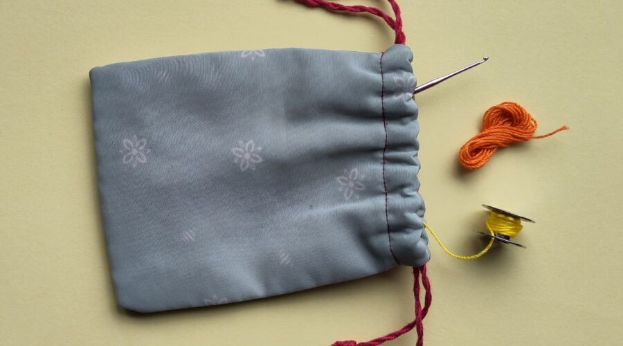 Ein liegendes, geöffnetes Stoffsäckchen aus dem eine Häkelnadel, eine Garnspule und ein Miniknäuel hervorschauen.