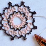 Ein symmetrisches Occhi-Spitzenornament in den Farben Rosa und Mokka, dezent mit PErlen verzeiert. In der rechten unteren Ecke ist ein Occhischiffchen abgebildet.
