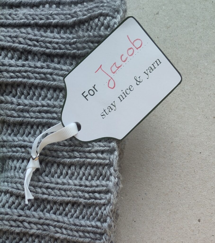 Ein Geschenkanhänger mit handschriftlich ergänztem Namen an einem Strickstück