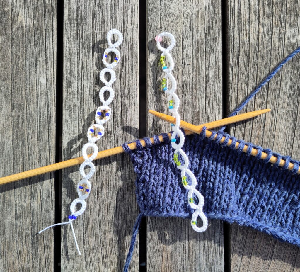 Ein eine blaue Strickarbeit auf Bambusnadeln mi t zwei weißen perlenverzierten Reihenzählen, die in Occhitechnik gefertigt wurden.