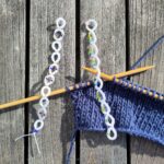 Ein eine blaue Strickarbeit auf Bambusnadeln mi t zwei weißen perlenverzierten Reihenzählen, die in Occhitechnik gefertigt wurden.