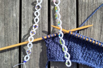 Ein eine blaue Strickarbeit auf Bambusnadeln mi t zwei weißen perlenverzierten Reihenzählen, die in Occhitechnik gefertigt wurden.
