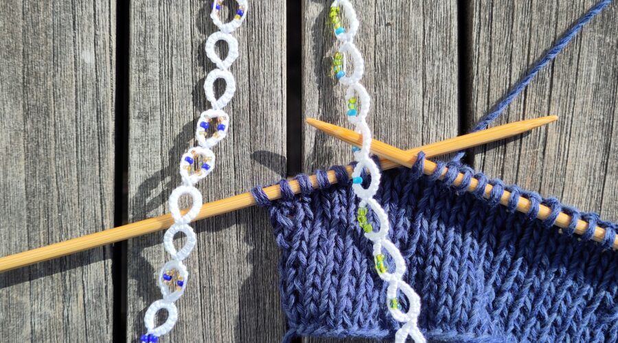 Ein eine blaue Strickarbeit auf Bambusnadeln mi t zwei weißen perlenverzierten Reihenzählen, die in Occhitechnik gefertigt wurden.